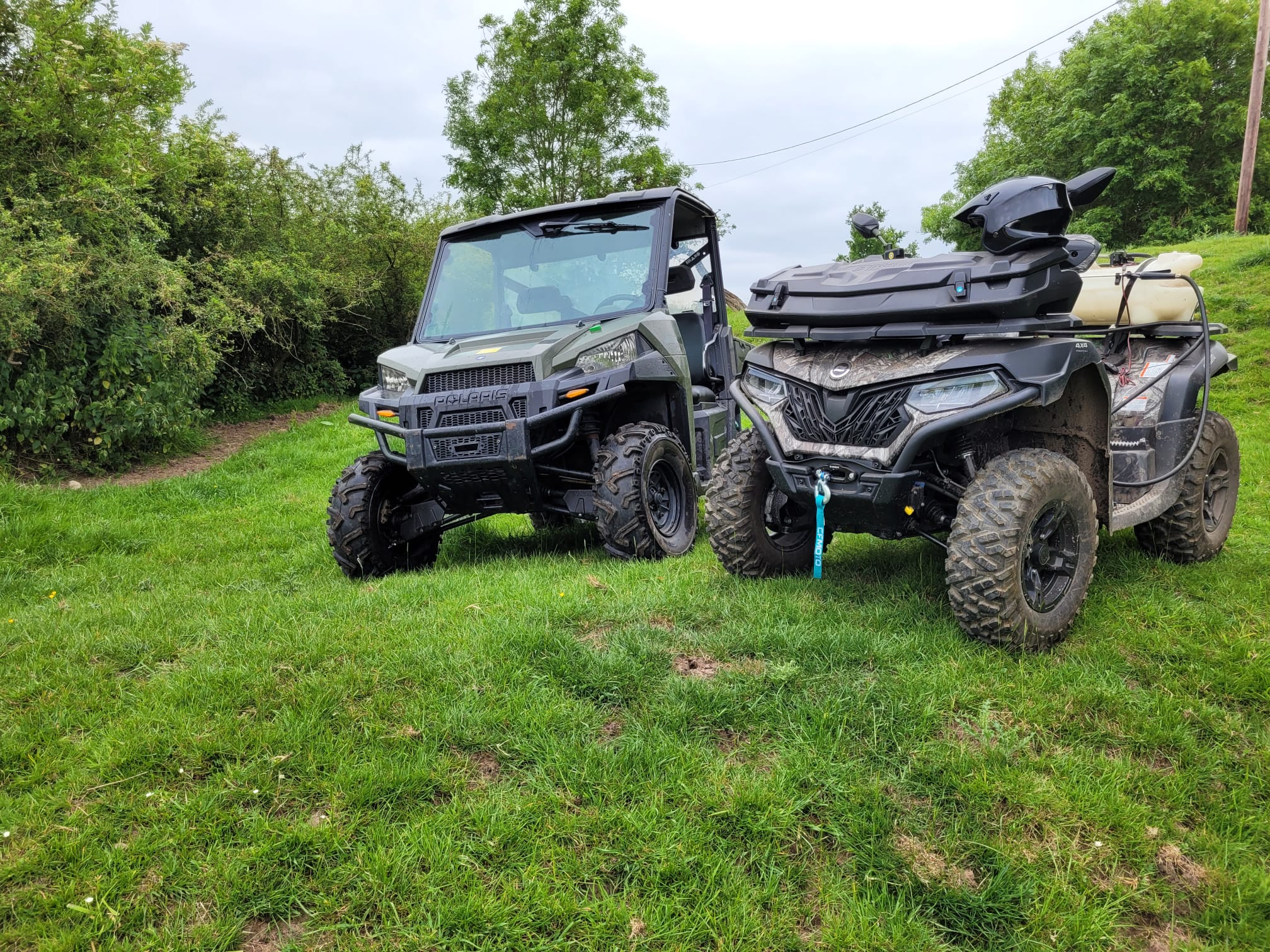 A Polaris utility terrain vehicle and a quad bike with front storage box parked side by side on grass beside trees. A Polaris utility terrain vehicle and a quad bike with front storage box parked side by side on grass beside trees.