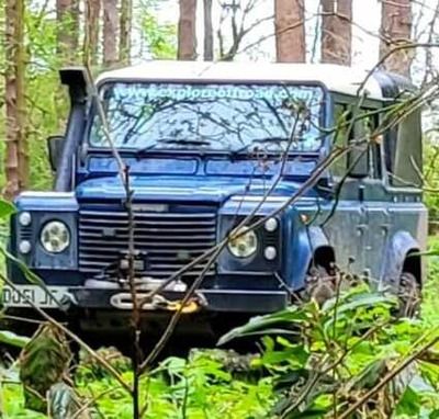 A blue Land Rover Defender with protective bars and recovery equipment among green woodland.