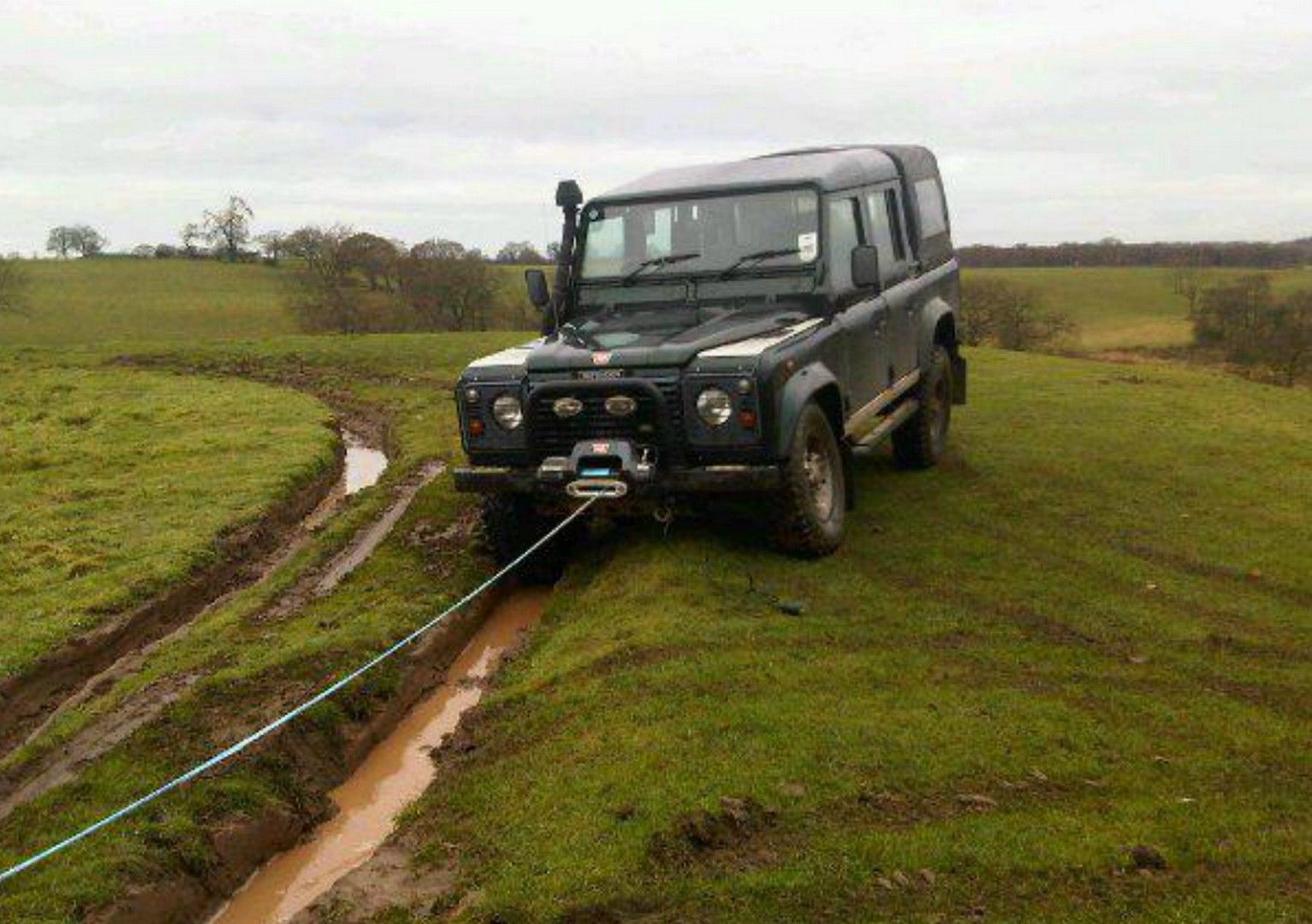 A grey Land Rover Defender on a grassy field with recovery lines extending from the front of the vehicle.